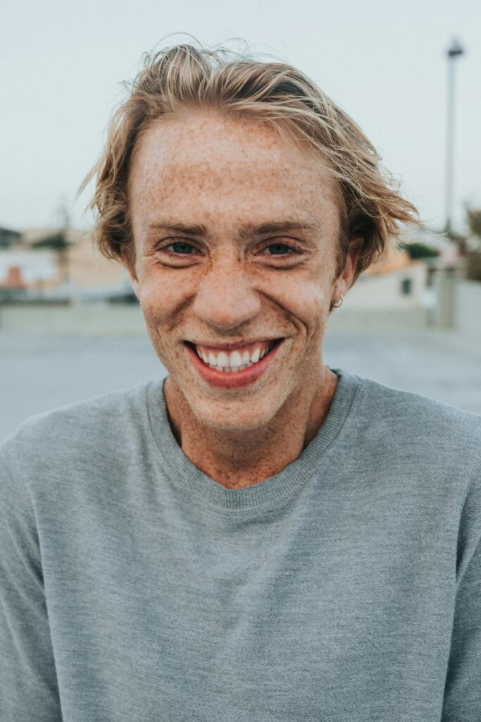 Close-up of a freckled man with blond hair smiling, wearing a gray sweater, outdoors.